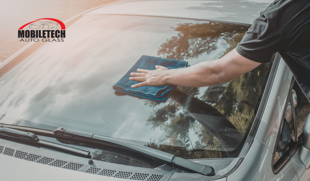 A Mobiletech Auto Glass technician wiping a car windshield with a blue microfiber cloth, with the company logo visible in the top left corner.