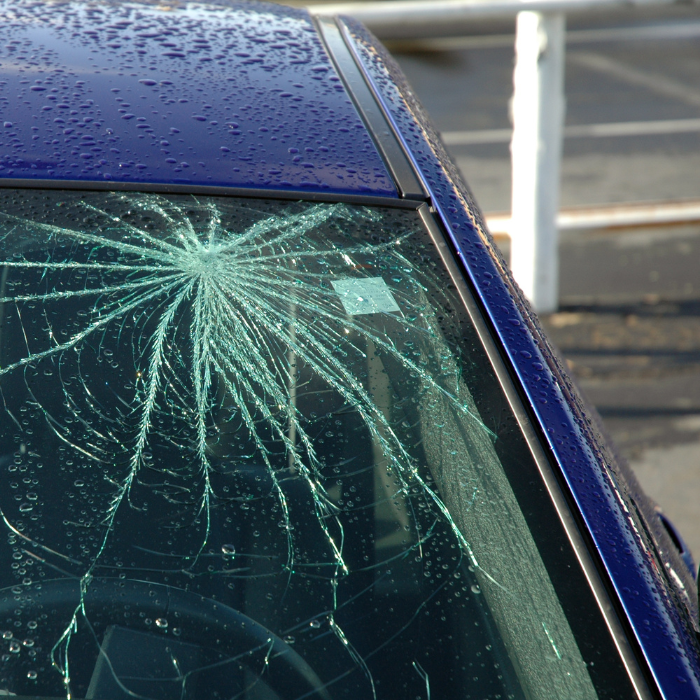 Close-up of a severely shattered windshield on a wet blue car, with a starburst crack pattern radiating from a central impact point, photographed outdoors after rain.