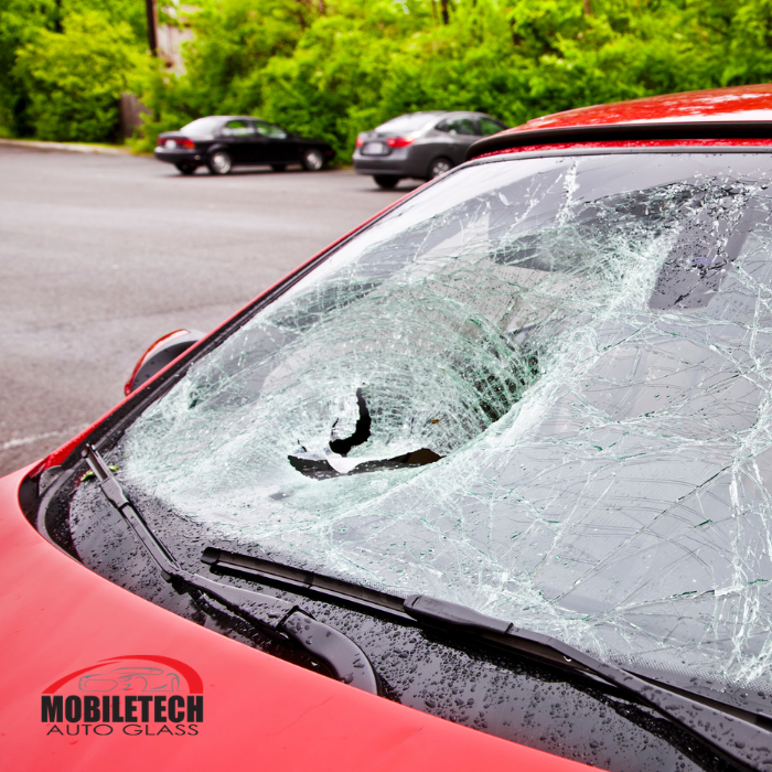 A Mobiletech Auto Glass promotional image showing a red car with a severely smashed and caved-in windshield in a wet parking lot, with the company logo in the bottom left corner.
