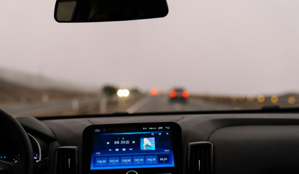 A view from inside a car dashboard showing a touchscreen infotainment system tuned to FM radio station 88.30, while driving on a wet, foggy highway with vehicles visible ahead through the windshield.