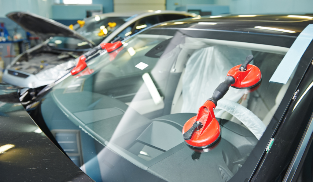 Red suction cup tools holding a replacement windshield in place on a black car during installation inside an auto glass repair shop, with another vehicle being serviced visible in the background.
